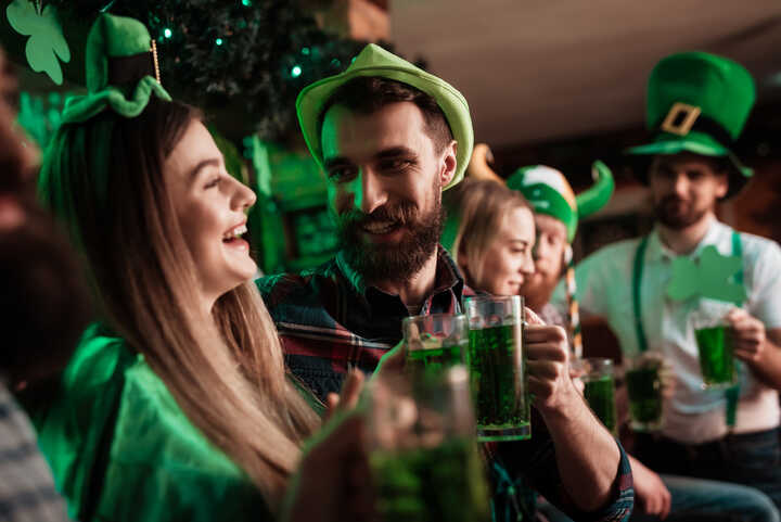People in a bar, smiling and cheersing glasses of stout in St. Patrick's Day hats, and green clothes