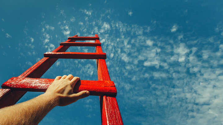 The top of a red painted ladder reaching into the sky with a hand on it, symbolising a challenge.