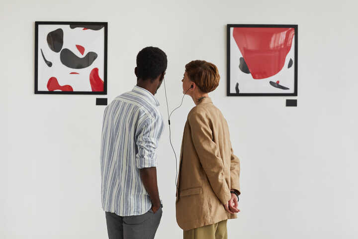A woman and man looking at art on the wall of an exhibit, while sharing a listening device to have it explained to them