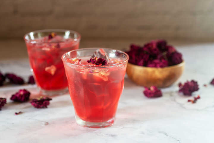 Two glasses of pink iced tea, with a bowl of rose petals in the background and rose petals intermittently scattered