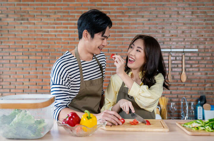 An Asian couple in cookery aprons. He's chopping strawberries, while she's feeding him one. They're both mid-giggle.