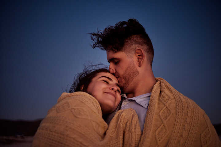 A couple wrapped in a cosy cable-knitted blanket at dusk. He is kissing her forehead as she smiles and looks at the sky