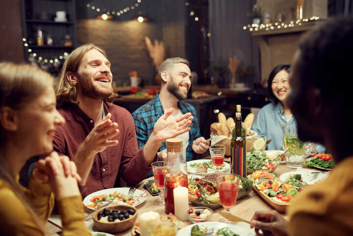 A group of friends laughing and enjoying a meal in a dimly-lit setting with fairylights in the background.