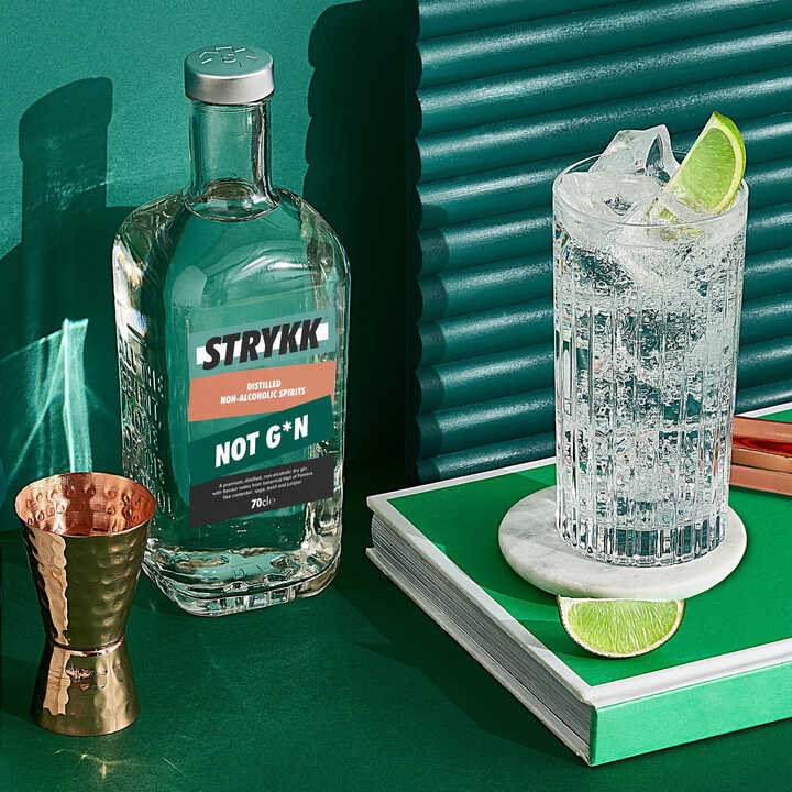 Bottle of Strykk Not Gin against a green background next to glasses with slices of lime in them
